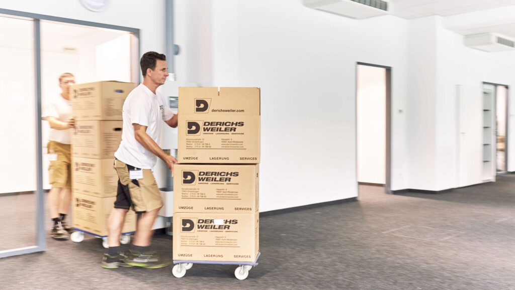 Two Derichsweiler employees move large cardboard boxes on a cart through an office hallway.