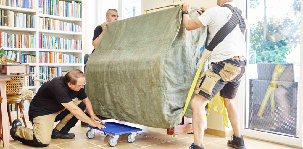 Drei Umzugshelfer transportieren ein großes, abgedecktes Möbelstück auf einer Transportkarre in einem hellen Raum mit Bücherregalen.