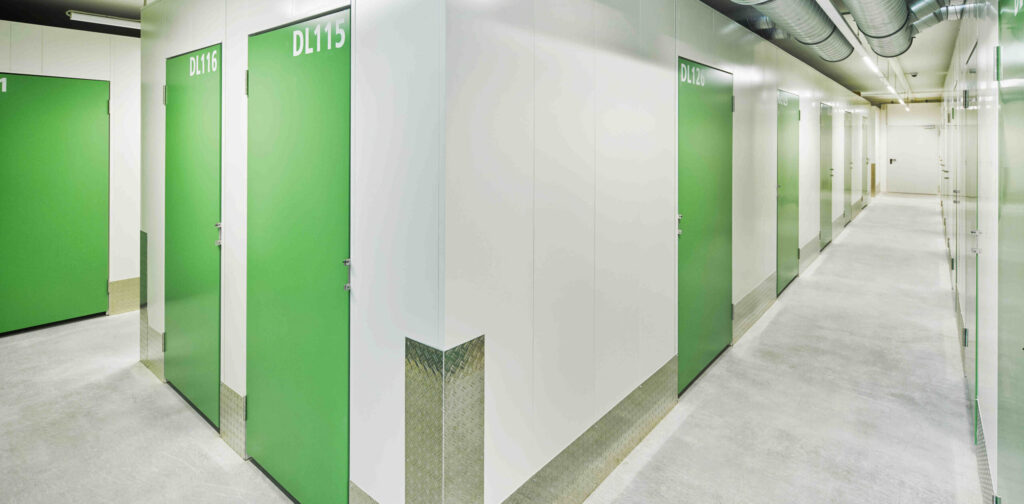 Ein hell beleuchteter Flur in einem Lagerhaus mit weißen Wänden und leuchtend grünen Türen, die mit Nummern beschriftet sind.