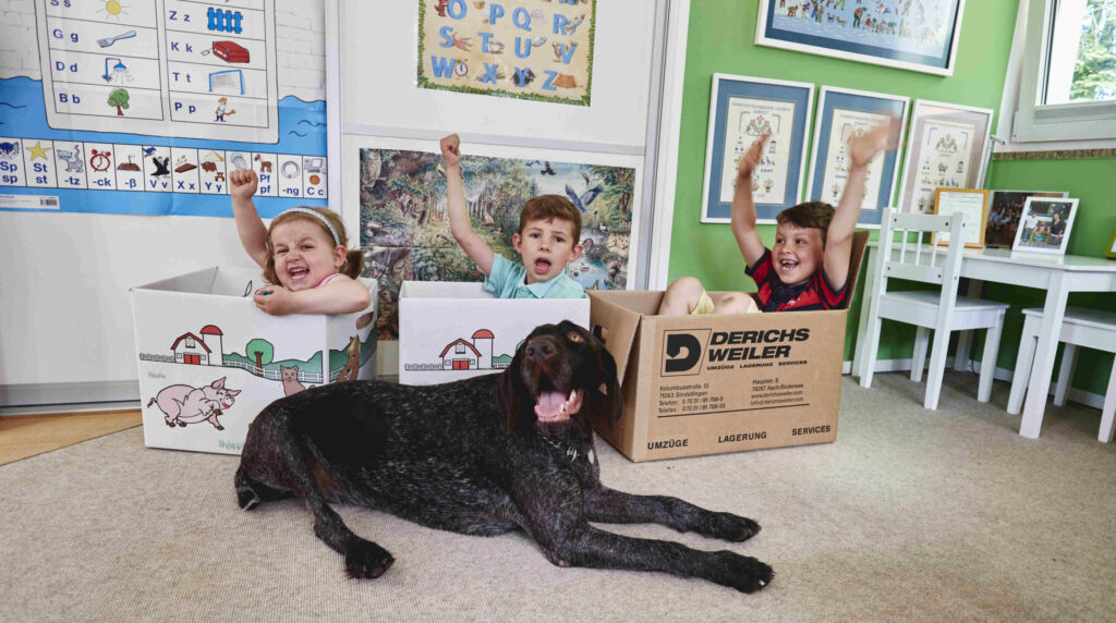 Drei Kinder sitzen in Umzugskartons in einem Schulklassenzimmer. Vor ihnen liegt ein schwarzer Hund.