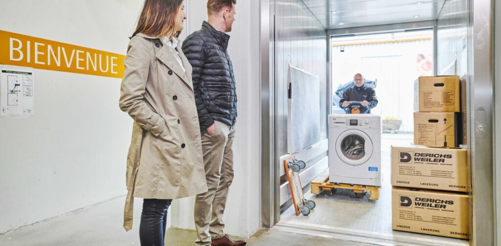 Ein Umzugshelfer schiebt eine Waschmaschine in einen mit Umzugskartons gefüllten Lastenaufzug. Zwei Personen stehen draußen.