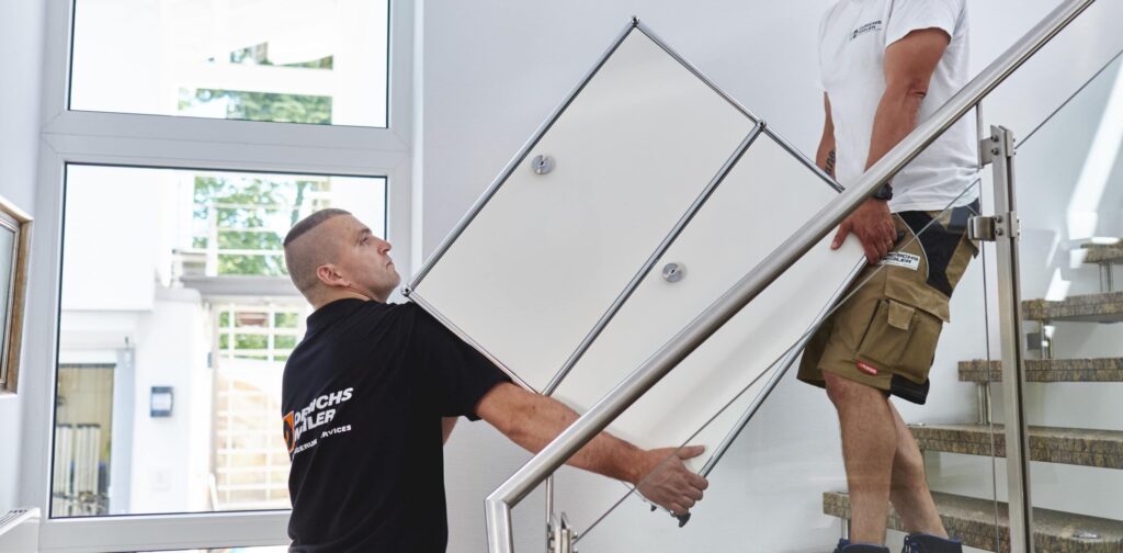 Zwei Männer tragen vorsichtig einen großen Schrank eine Treppe hinauf in einem hellen, modernen Haus.