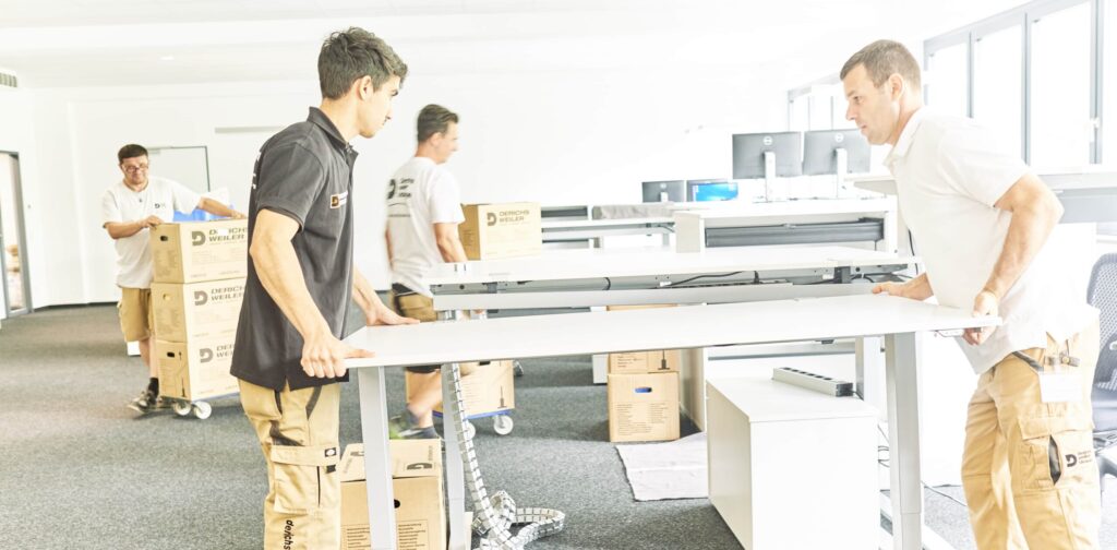 In einem hellen Büro montieren Arbeiter Tische und transportieren Kartons.