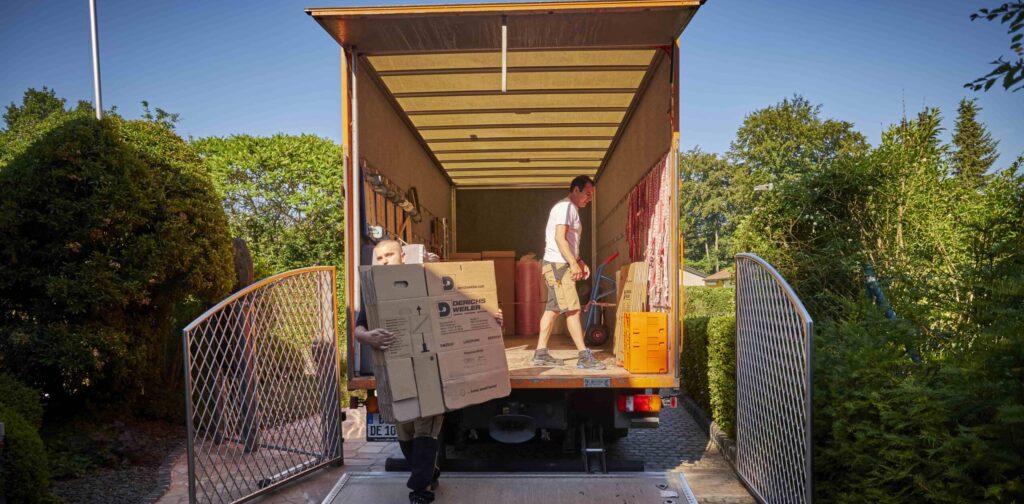 An einem sonnigen Tag tragen zwei Personen Kisten aus einem offenen Umzugswagen.
