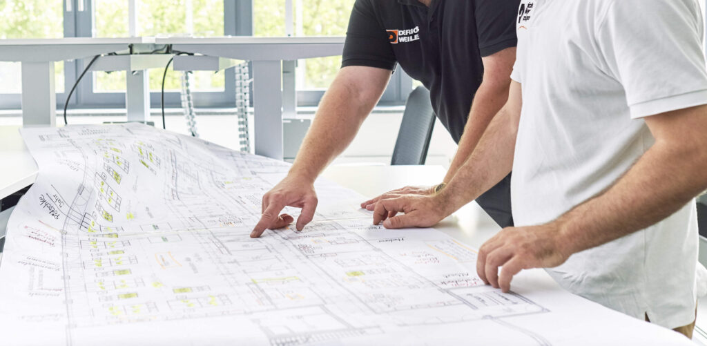 Zwei Männer in einem hellen Büro betrachten einen großen Bauplan auf einem Tisch.