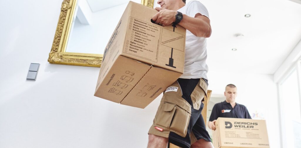 Zwei Umzugshelfer tragen Kartons in einem hellen Raum mit einem goldgerahmten Spiegel an der Wand.