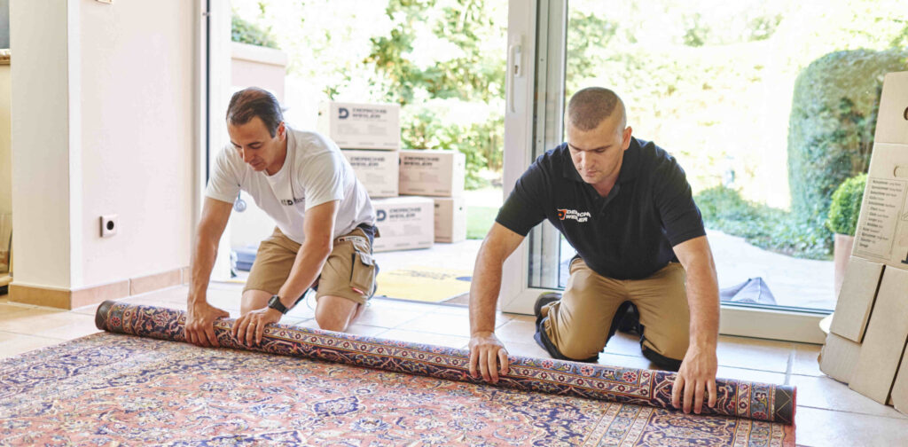 Zwei Umzugshelfer knien auf einem Fliesenboden und rollen vorsichtig einen gemusterten Teppich auf.